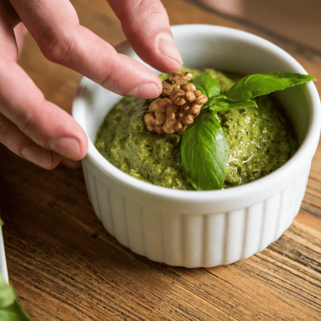 Homemade pesto recipe without pine nuts in a glass jar, surrounded by fresh basil, garlic, and parmesan.