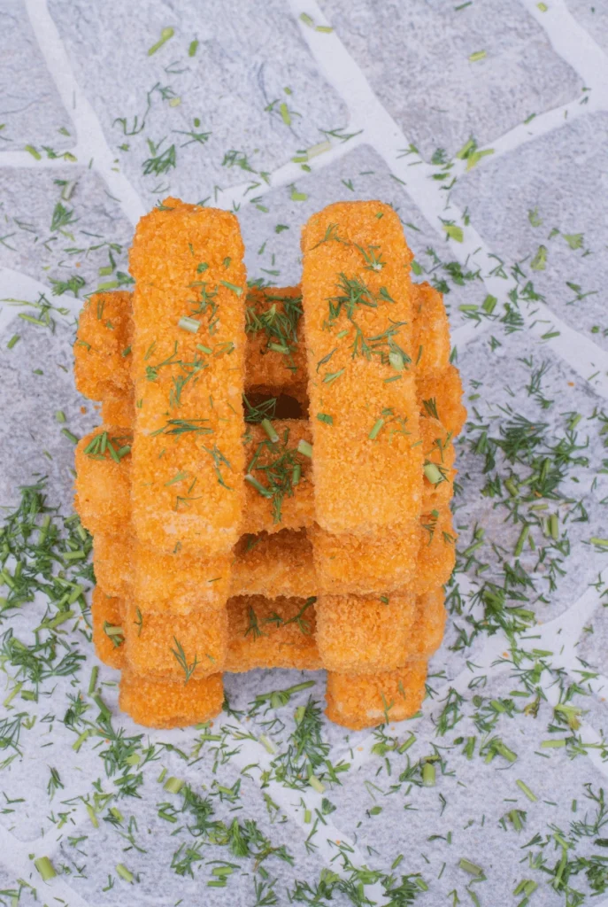 How to Make Pizza Crunchers That Everyone Will Love 2 A stack of breaded fish sticks arranged in a crisscross tower pattern, garnished with chopped dill on a light stone surface.